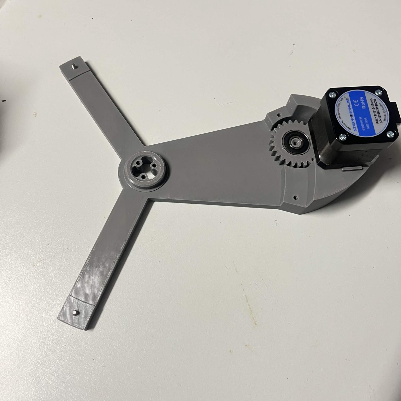 The three-armed grey NEMA bracket seen from above, with the 24-tooth idler gear sitting on its post with the 608 bearing uppermost, the stepper motor bolted to the outer end of the bracket, and the raised hub at the centre of the bracket