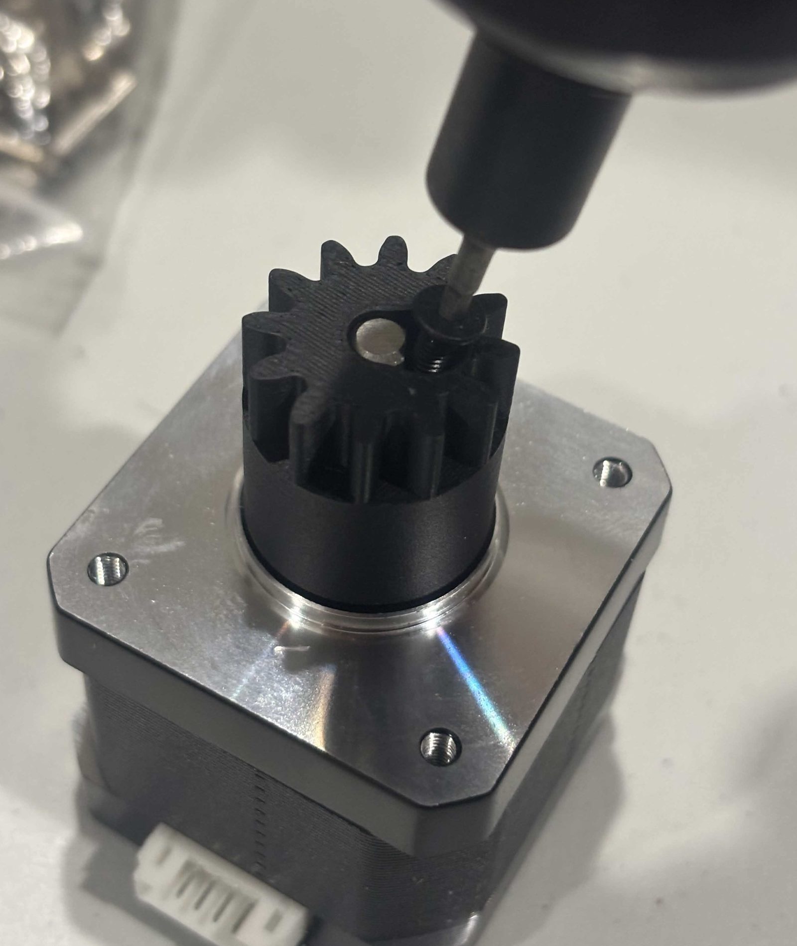 The charcoal 12-tooth input gear pushed onto the shaft of a NEMA 17, seen from above. A driver bit is turning the clamping screw, which stands in a hole in the end face beside the bore with its thread still showing. The flat on the motor shaft is visible through the bore next to it