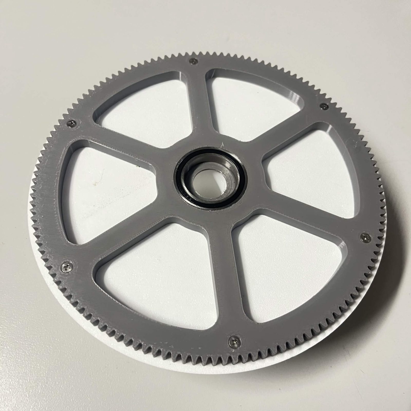 The grey 130-tooth output gear with a black-sealed 6806 bearing pressed into its centre, bolted onto a white rotor behind it, with a countersunk screw at the outer end of each of the six spokes