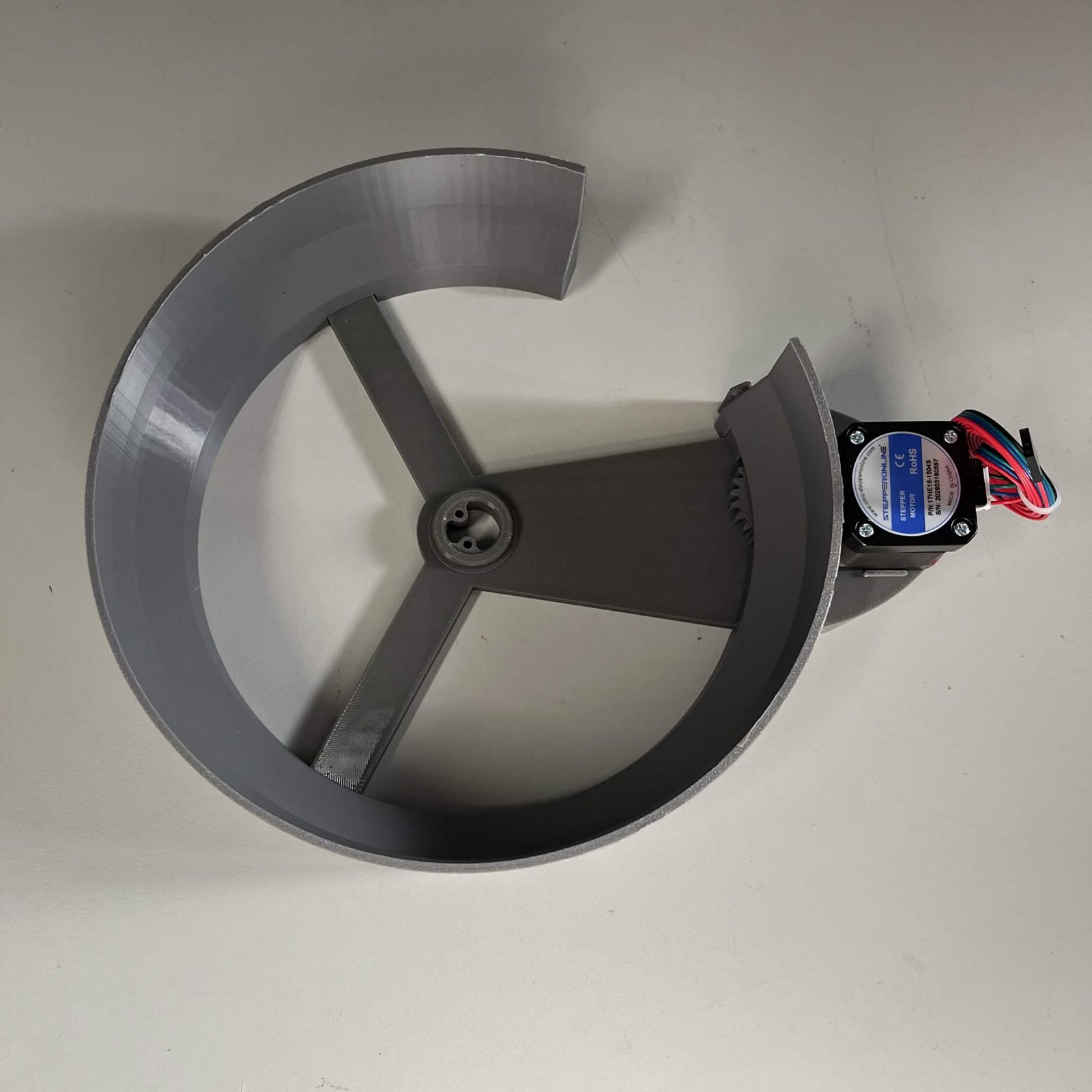 A finished channel core from directly above: the stator ring open at one side, the three-armed NEMA bracket fastened across it with the raised hub at its centre, the gear train at the ring's edge and the stepper motor standing outside the wall with its lead, and no rotor in the hub