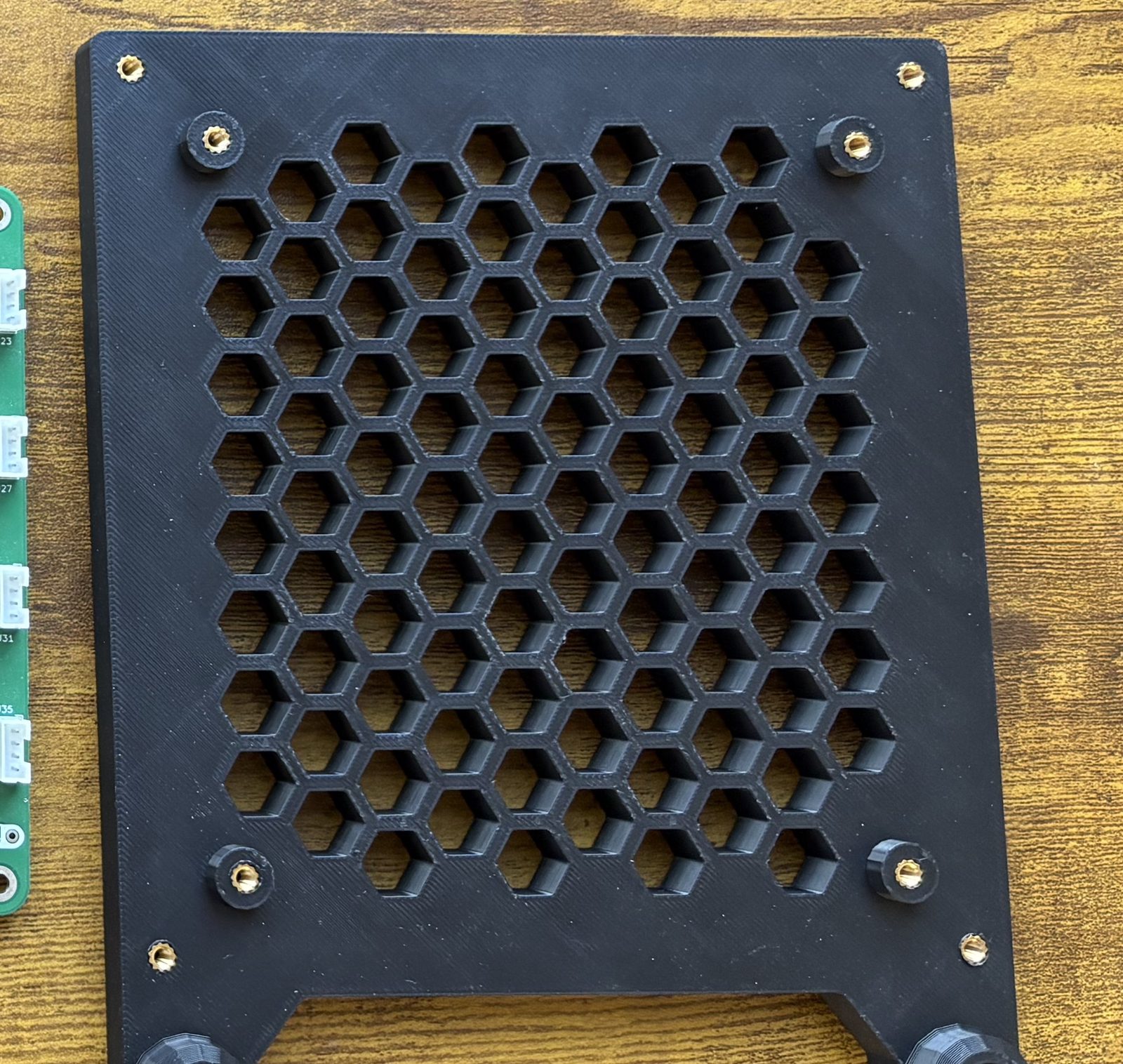 The printed housing base seen from above, showing the large honeycomb vent in its floor and eight brass M3 heat inserts, four on raised bosses inside and four at the outer corners