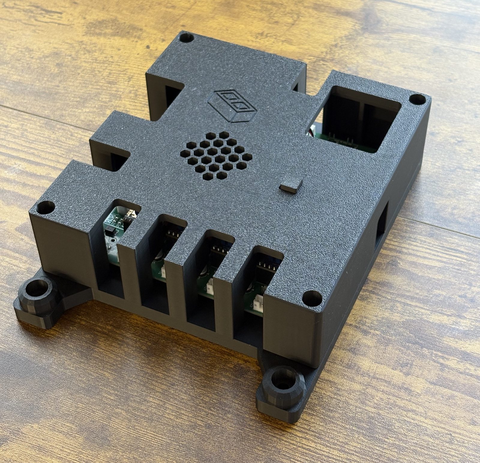 The finished housing at an angle, showing the honeycomb vent and basically logo on the lid, the square plunger head in the lid surface, and the bays along the right edge that expose the stepper connectors