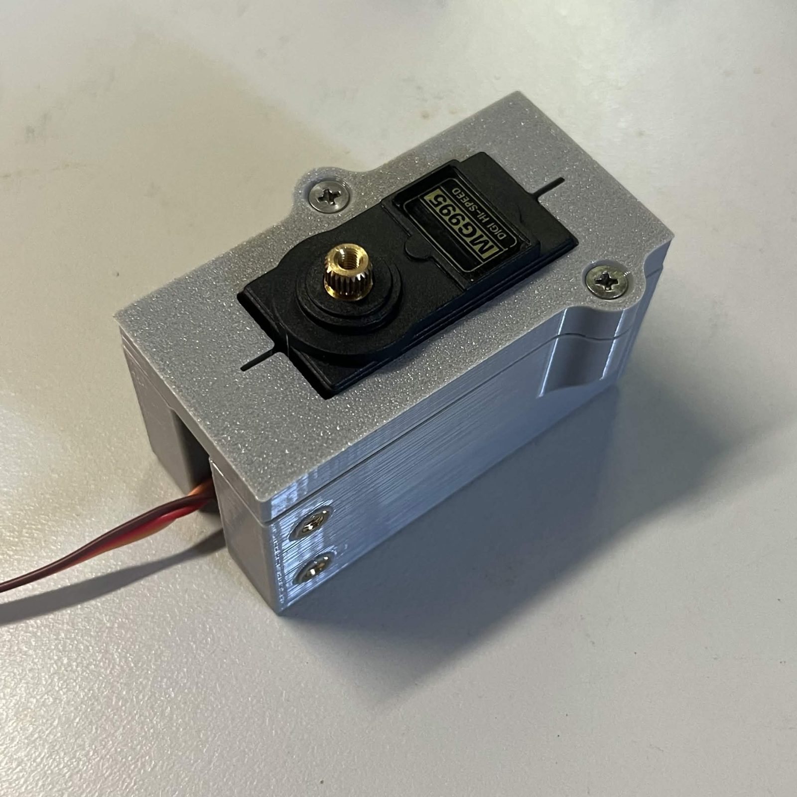 The same housing from the corner with the cover screwed down over the servo, two countersunk screw heads in the cover and the servo's splined output standing through the window in it
