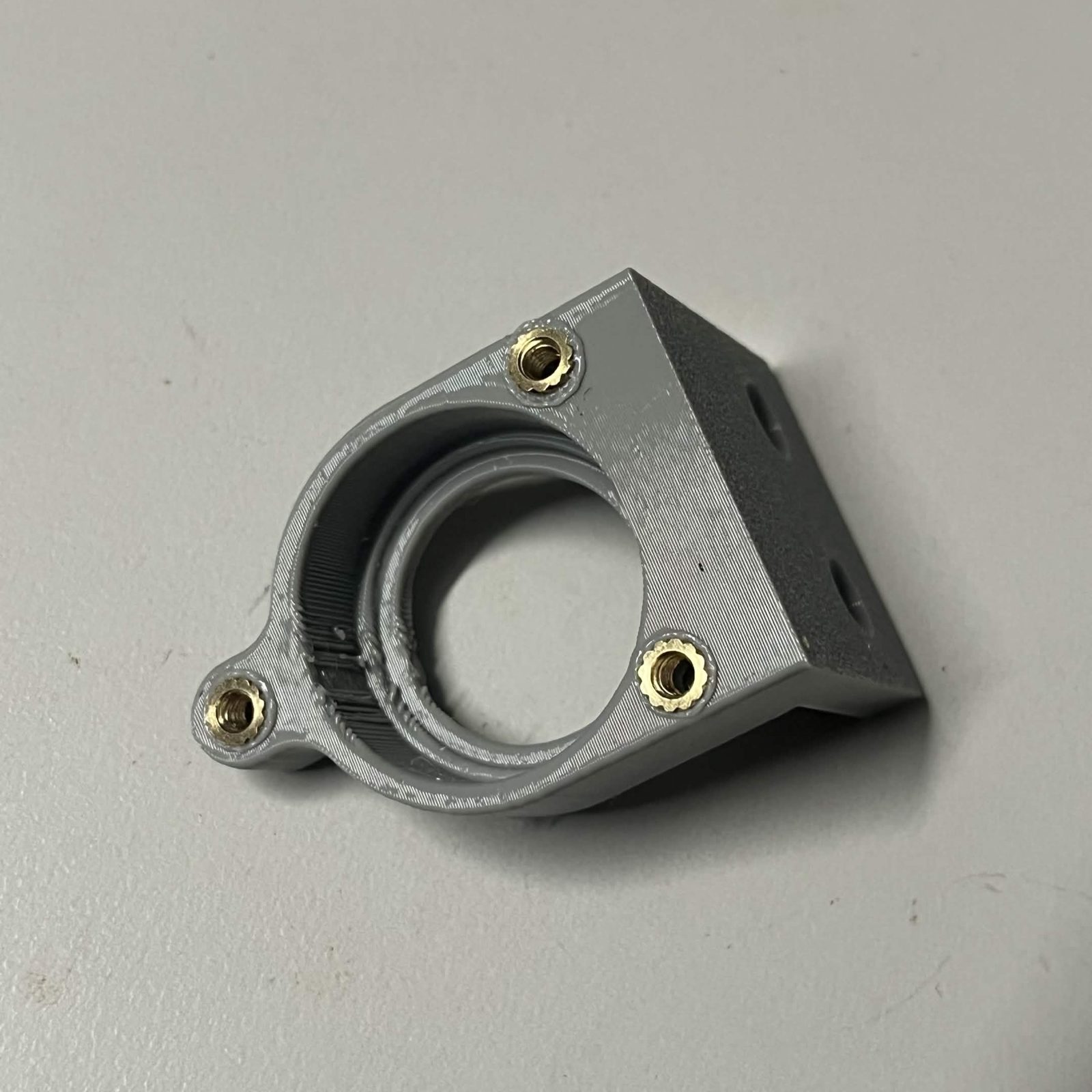 A printed bearing holder held at an angle, three brass heat inserts spaced around its empty bearing bore, one on the small ear and two on the body