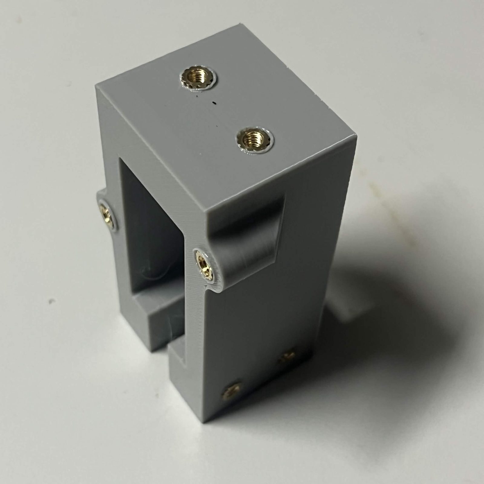 A printed servo bracket housing standing on end, brass heat inserts pressed into all six pockets: two on the top face, two on the front face beside the open pocket, two on the bottom edge