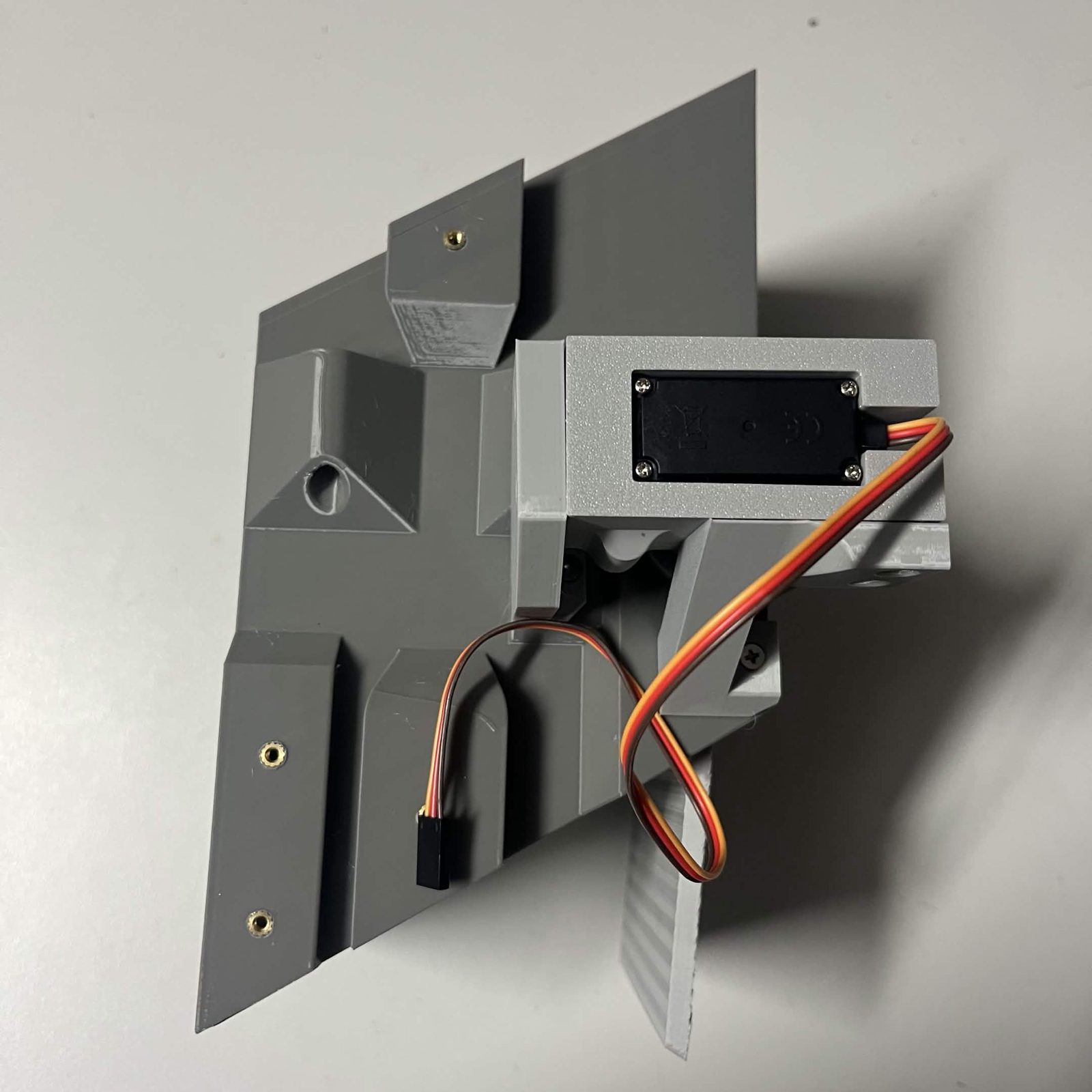 A finished door module bolted to a chute core: the MG995 in its printed bracket with its lead looped out to a bare connector, the bracket's arms screwed to the core's side, and the light grey door plate hanging below the core