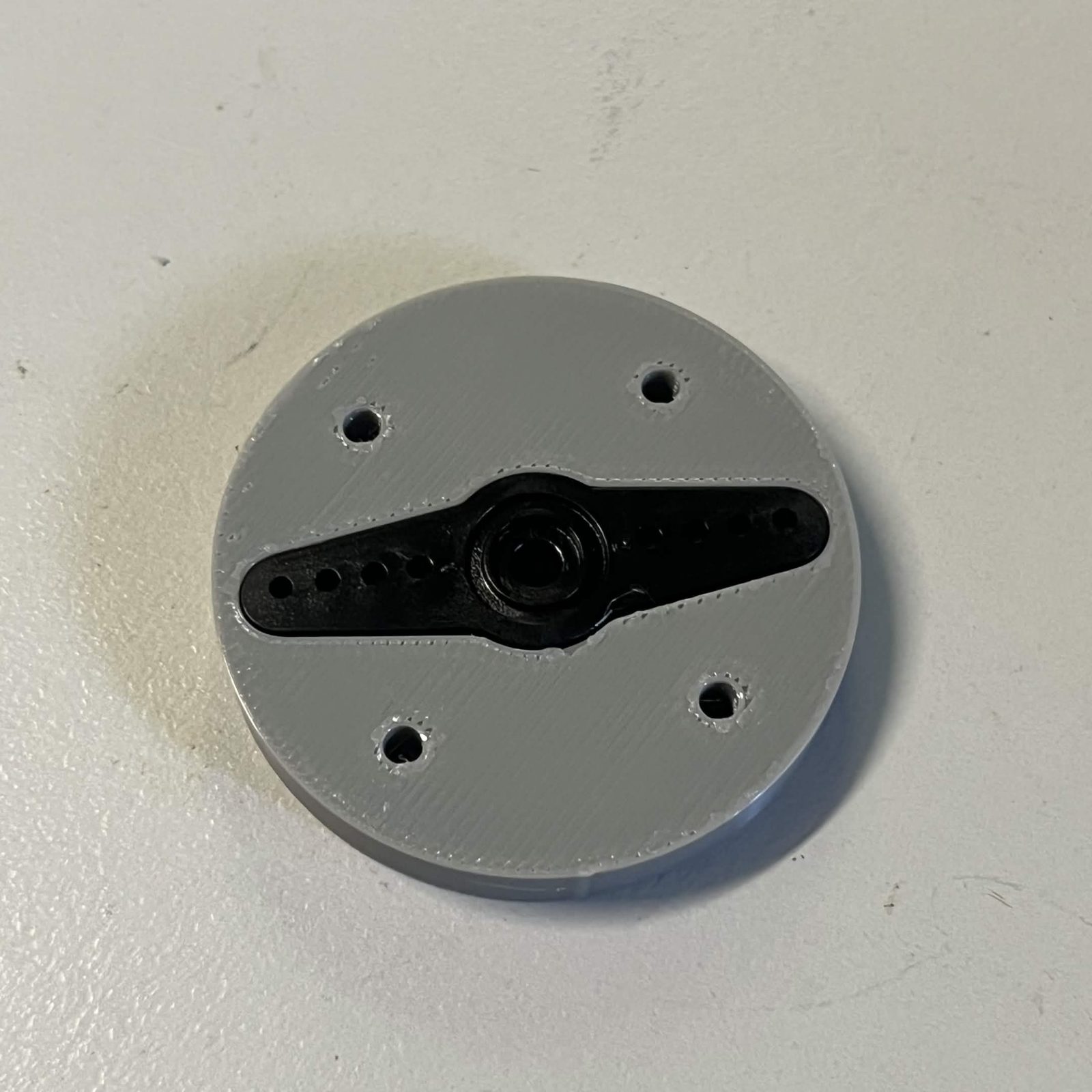 The finished servo adapter seen from the servo side: the black two-arm horn lying flush in its slot in the grey printed disc, the splined sleeve's opening in the middle of it and four empty countersunk holes around the face