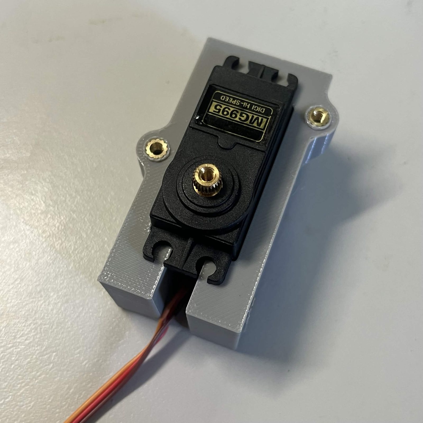 An MG995 servo lying in the open pocket of the printed housing with the cover off, its mounting tabs resting across the two brass-lined lugs and its lead running out of the bottom of the pocket