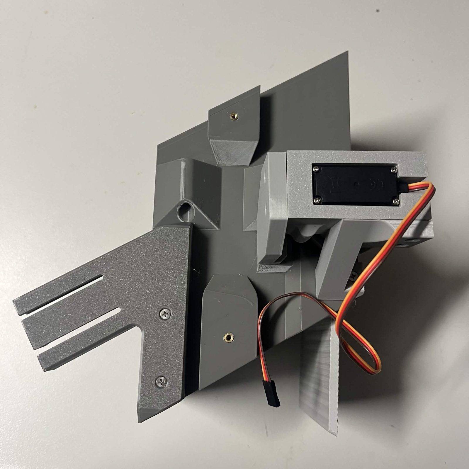 A funnel bracket bolted to the side of a chute core on the bench, its two countersunk screws driven home in the face and its three slotted arms standing clear, with the servo bracket and the door at the other end of the core