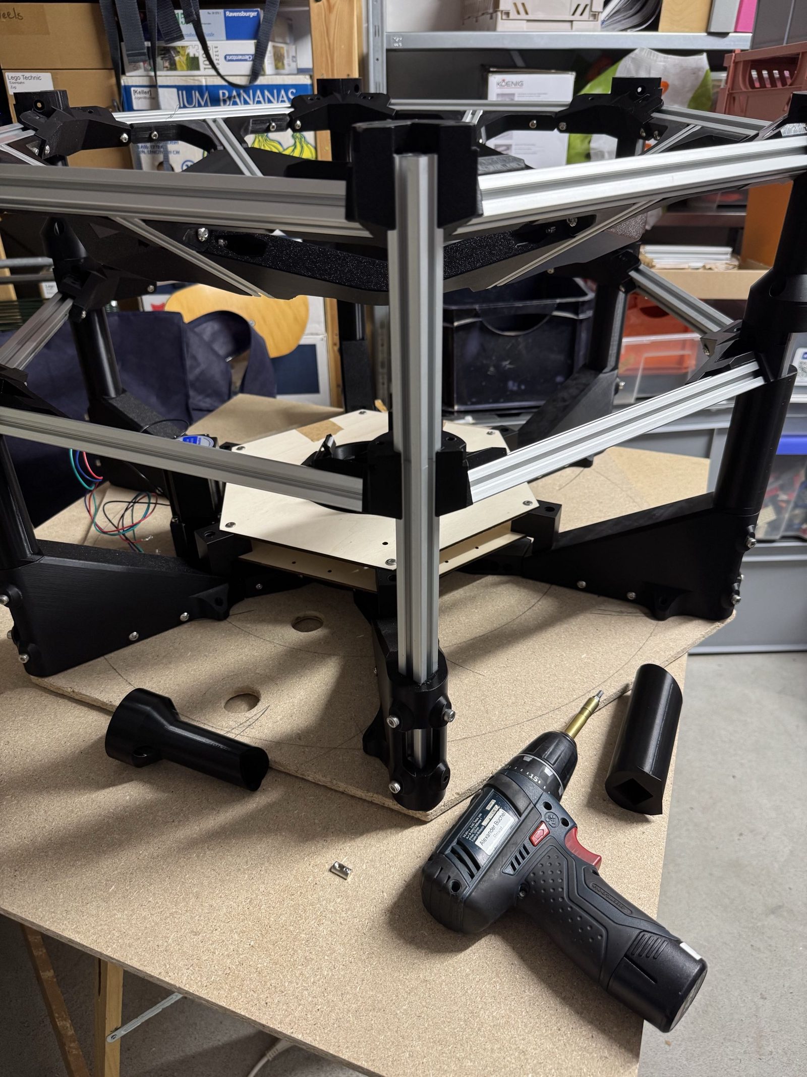 A partly built machine standing upside down on a workbench, resting on its top plate, with the interface's vertical extrusions pointing up and a hex frame layer being added on top of them, a cordless driver on the bench beside it