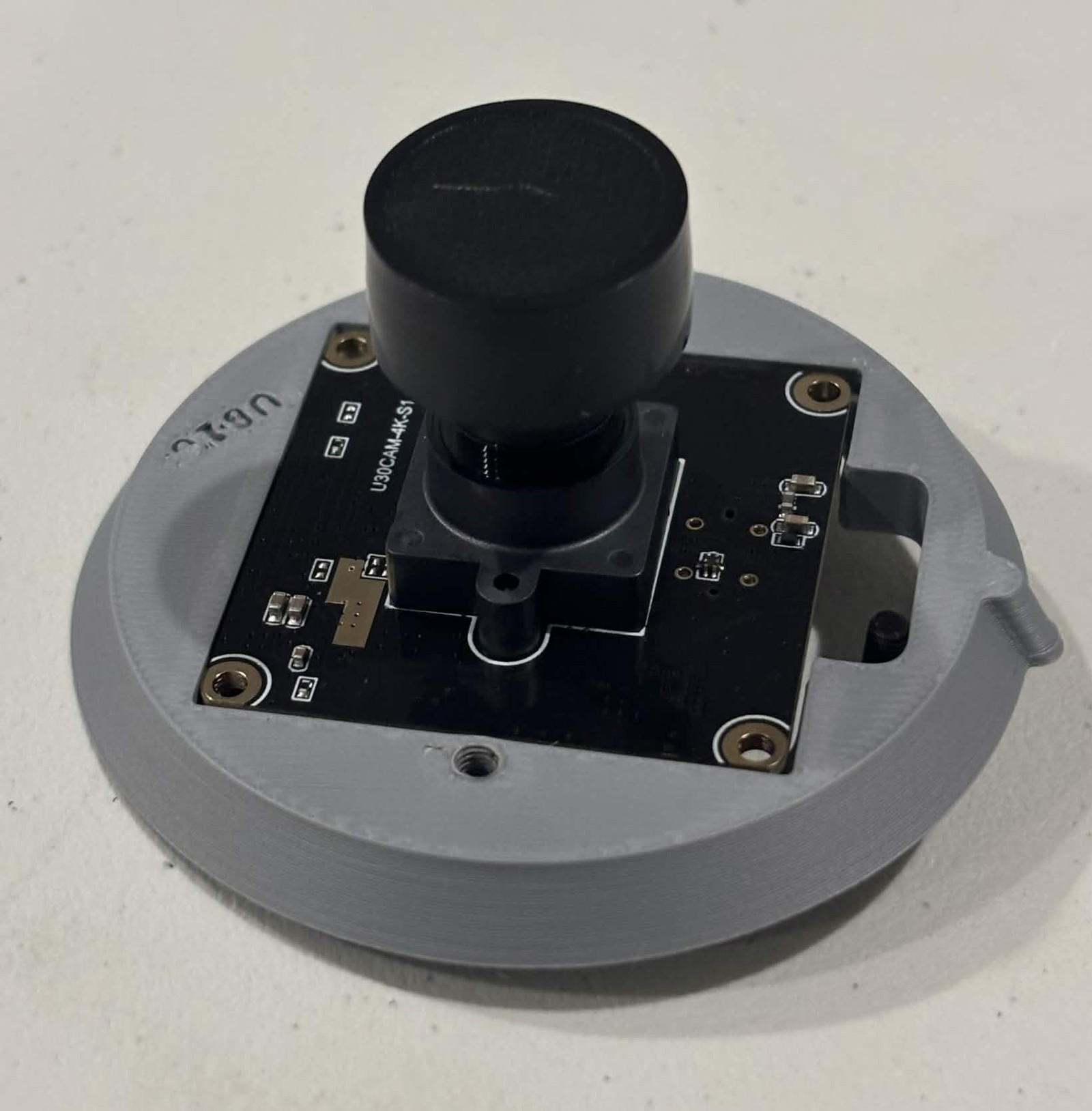 A camera board sitting in the square recess of a grey printed clasp half, component side up with the black cylindrical lens standing in the middle, and a screw hole in the plastic below the board