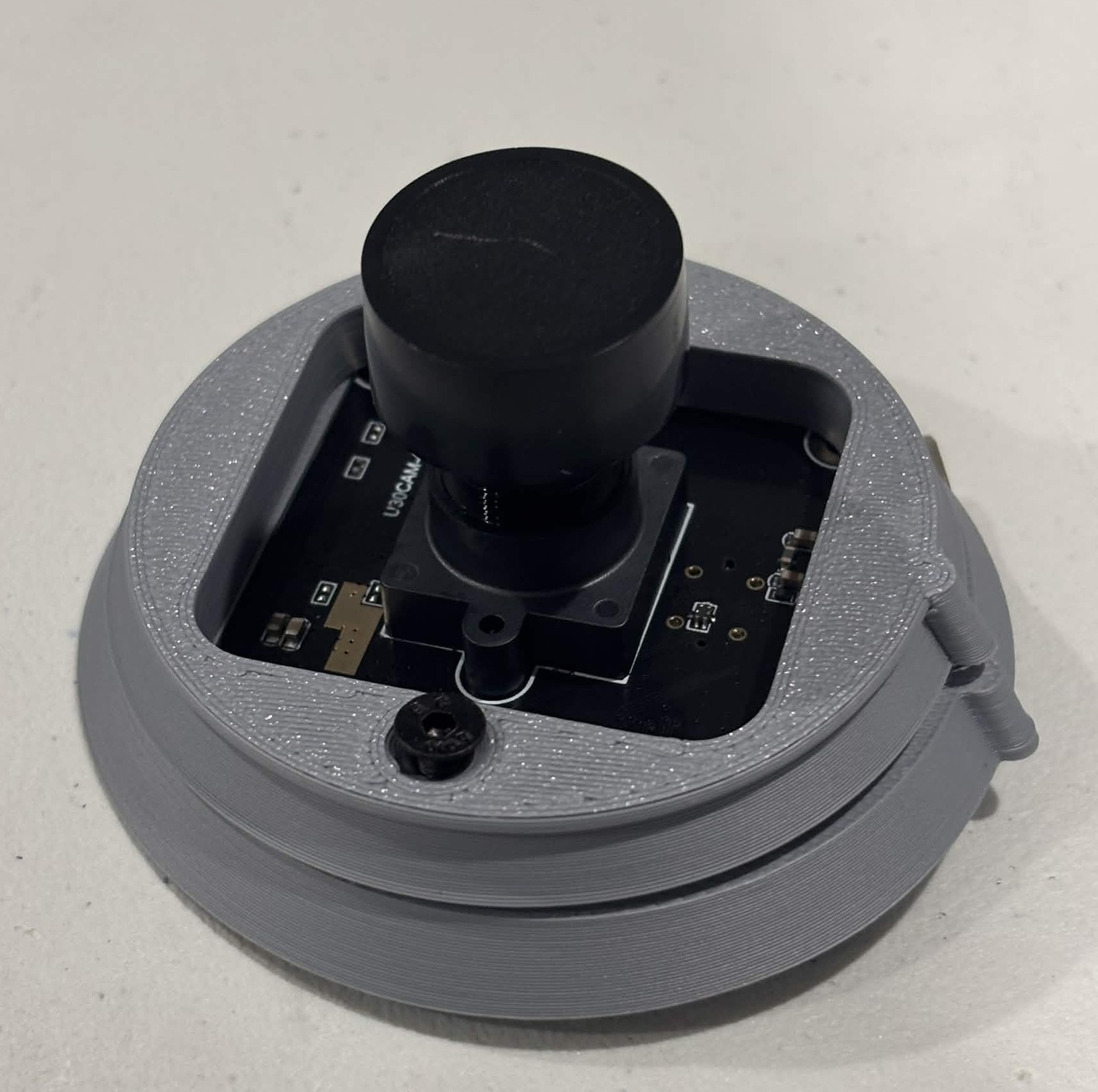 The second clasp half closed over the camera board, the lens standing through the square opening in it and a black countersunk screw seated flush in the plastic at the near edge