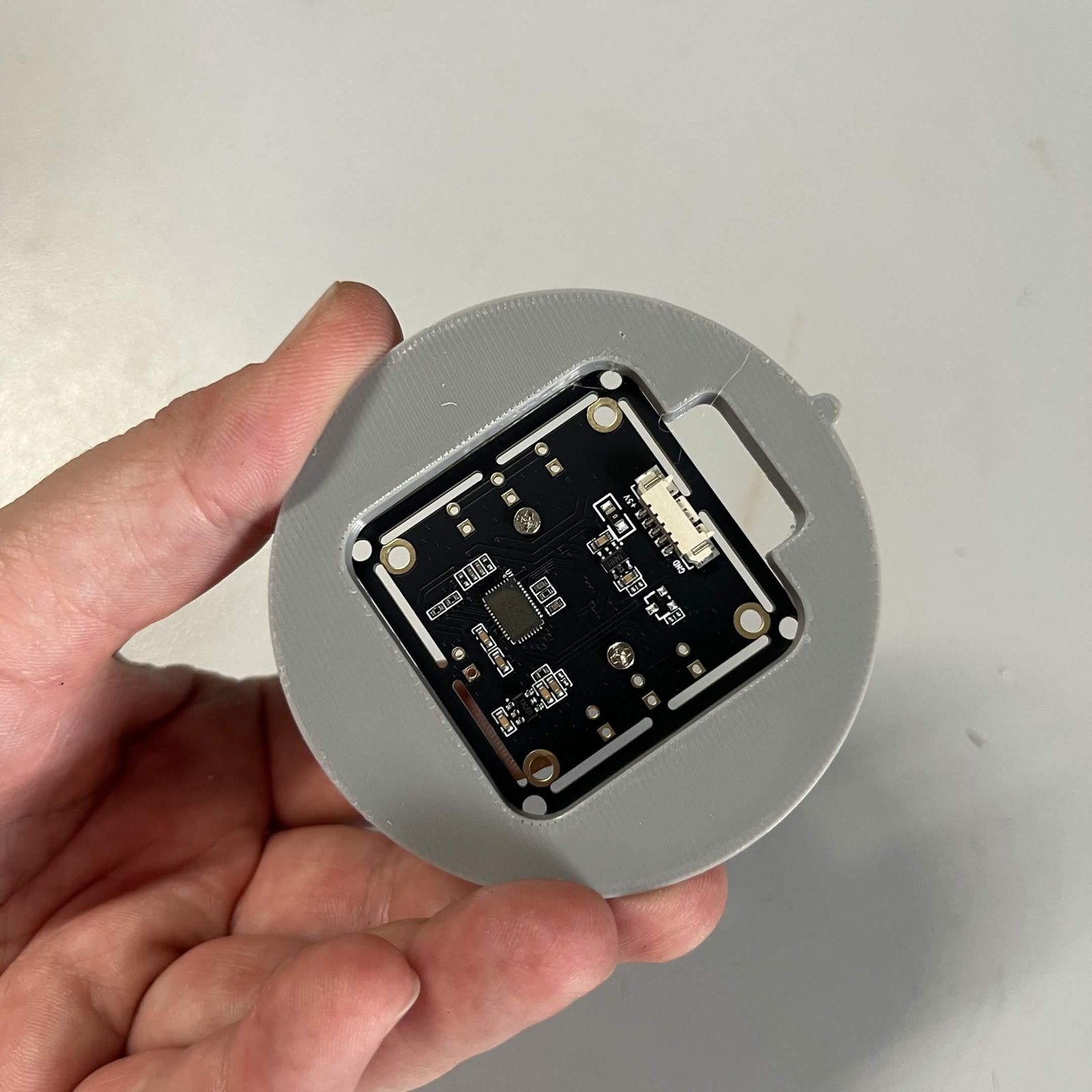 A hand holding the closed camera clasp, the camera board seated in the round grey disc with its ribbon socket empty and a rectangular slot in the plastic beside it