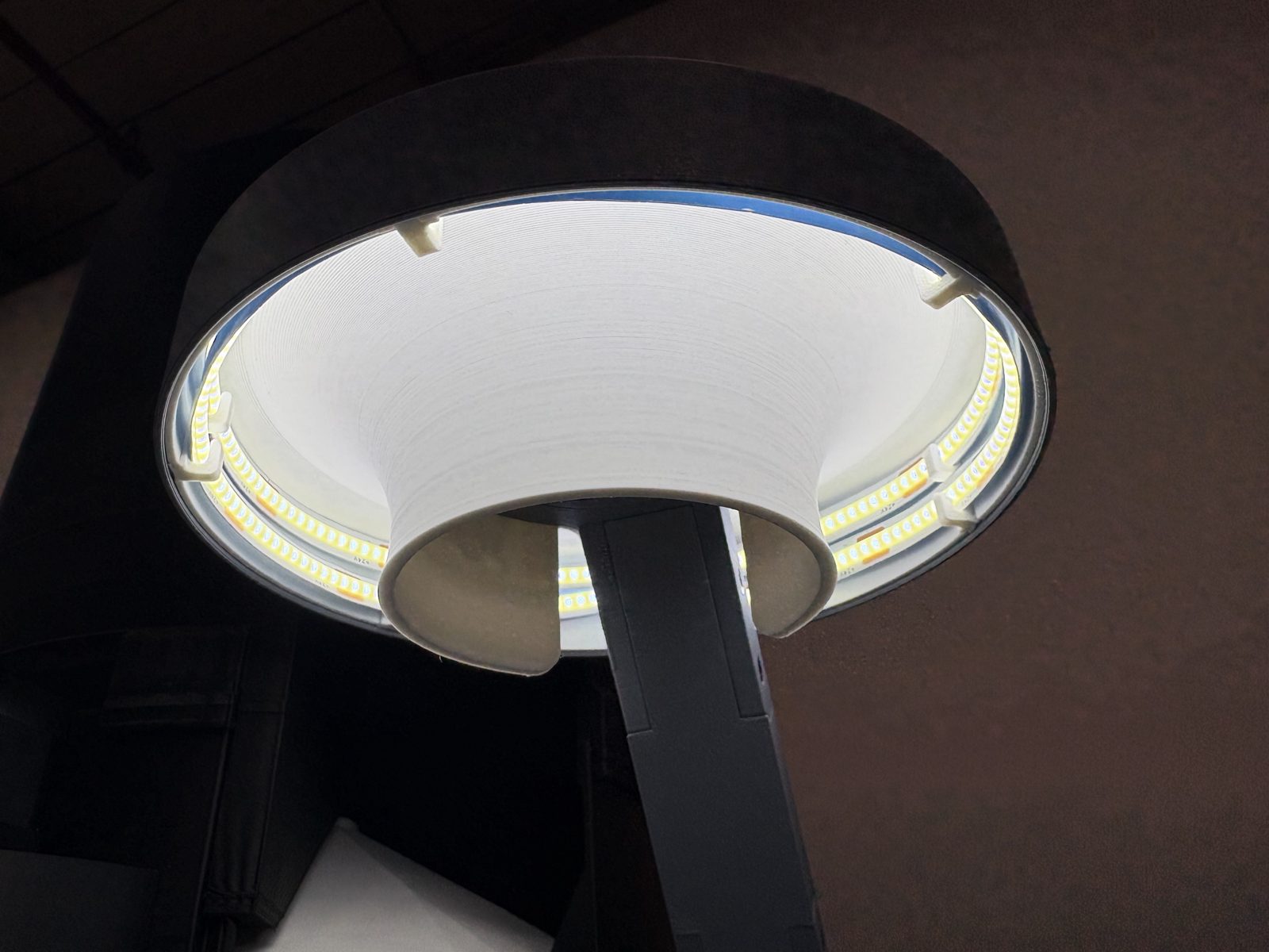 The lamp lit, photographed from underneath: a ring of LED strip glowing around the outside of the white reflector, the reflector's central funnel in the middle, and the arm behind it