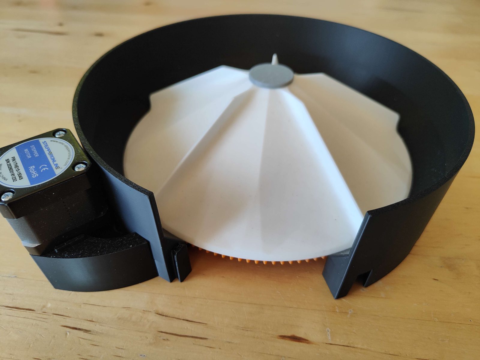 A finished classification channel on a wooden bench: the white finned rotor sitting down in the black stator ring with a small grey cap at its centre, the stepper motor at the left and the orange output gear showing at the gap in the ring