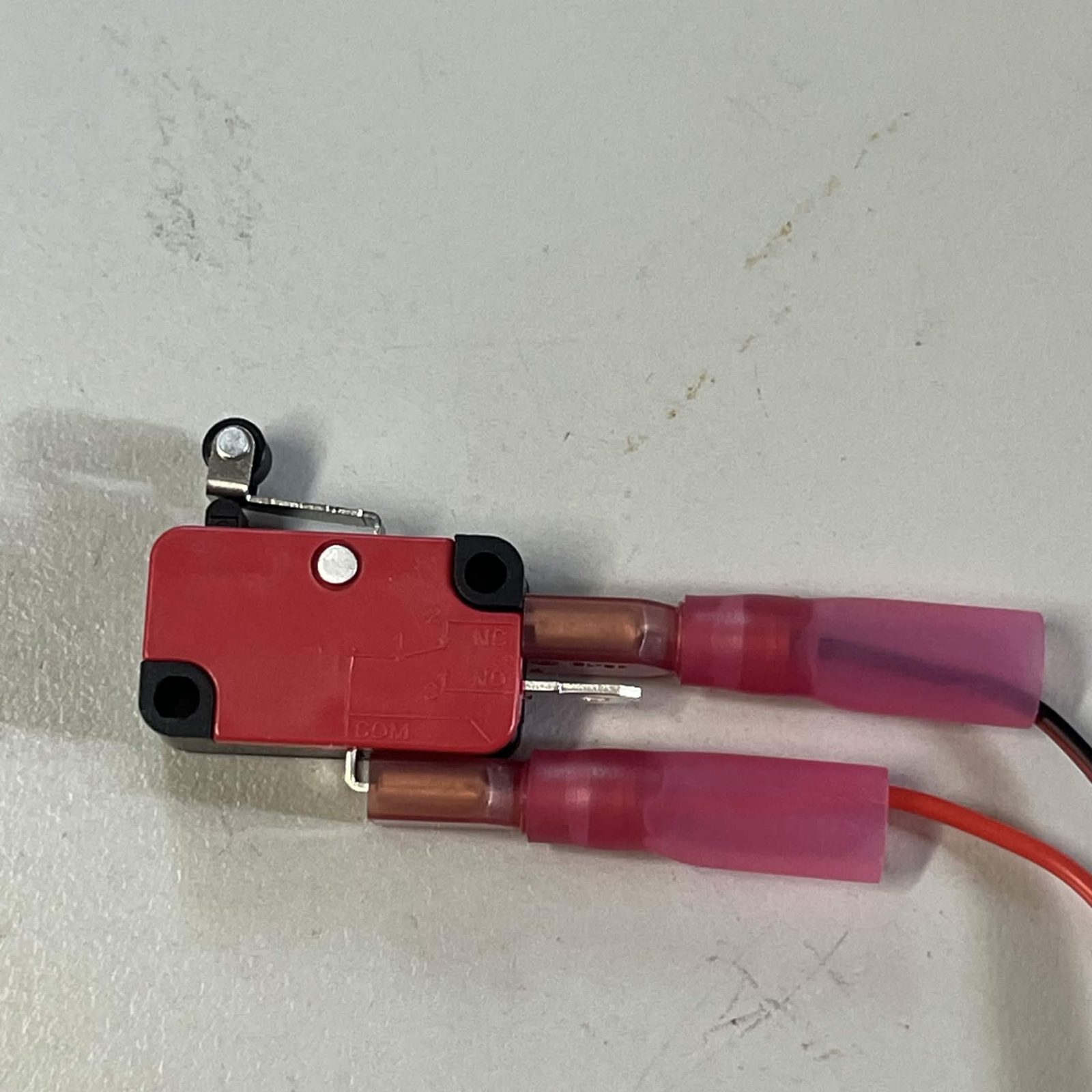 The red roller-lever switch on a bench with its printed schematic visible, NC and NO labelled against the two tabs on its side and COM against the tab on its bottom edge, an insulated receptacle pushed onto the NC tab and another onto the COM tab, and the middle NO tab left bare