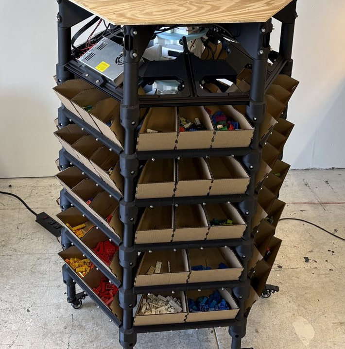 A bin tower on castors under its plywood deck, five layers of folded cardboard bins with sorted LEGO in them