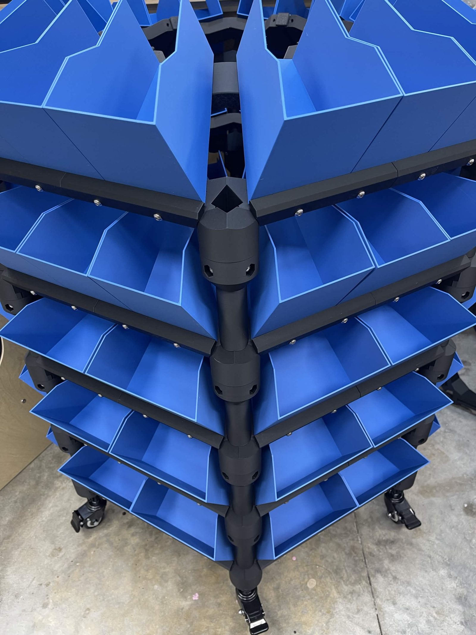 A loaded distribution stack seen from above and to one side, five layers of blue printed bins radiating out around the corner column, casters on the floor below