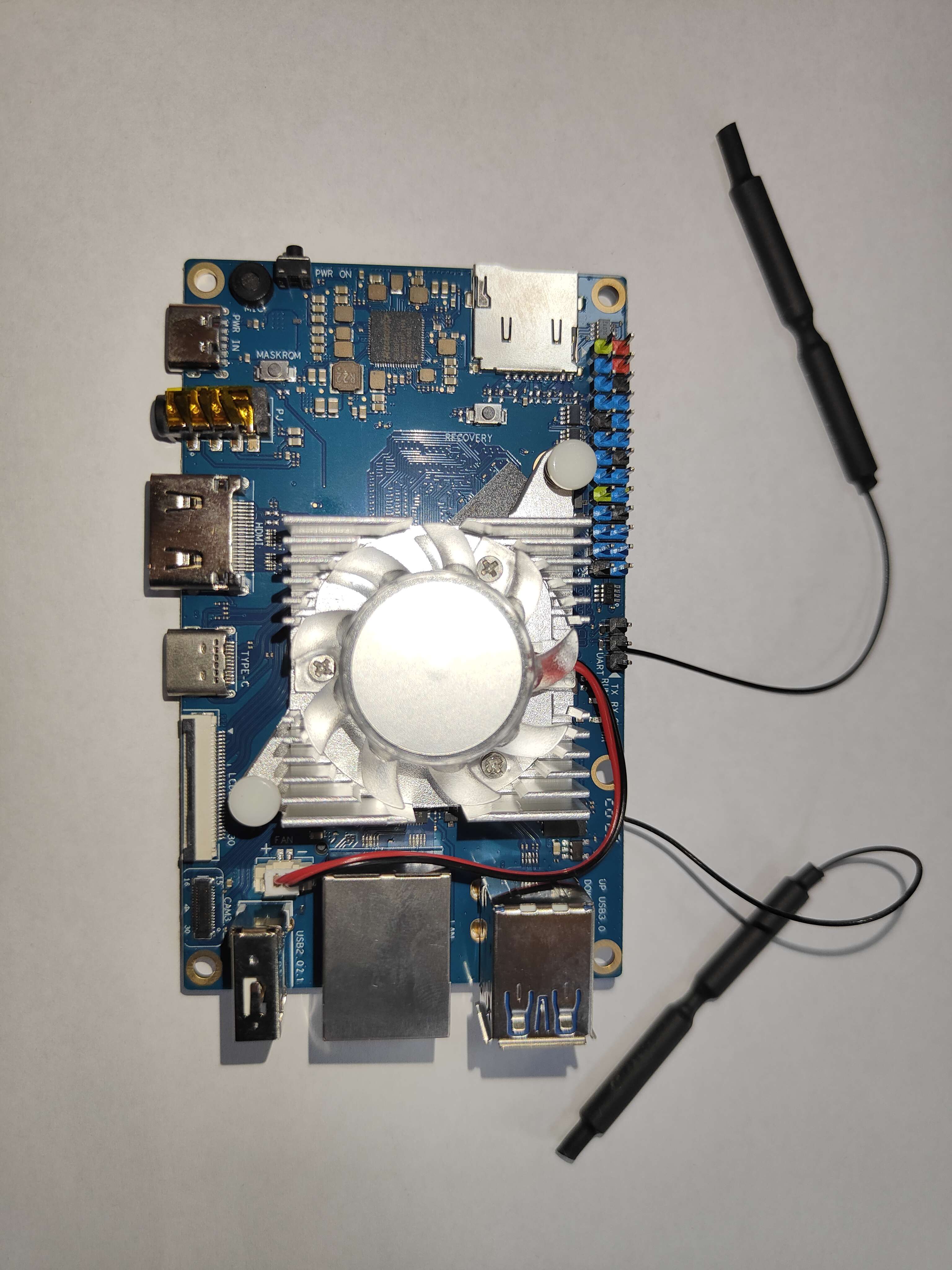 An Orange Pi 5 seen from above with the finned heatsink fan fitted over the SoC in the middle of the board, a white spring pin clipped through the board at opposite corners of the heatsink, the red and black fan lead running to the small white FAN socket below it, and two antenna leads trailing off the right-hand edge of the board