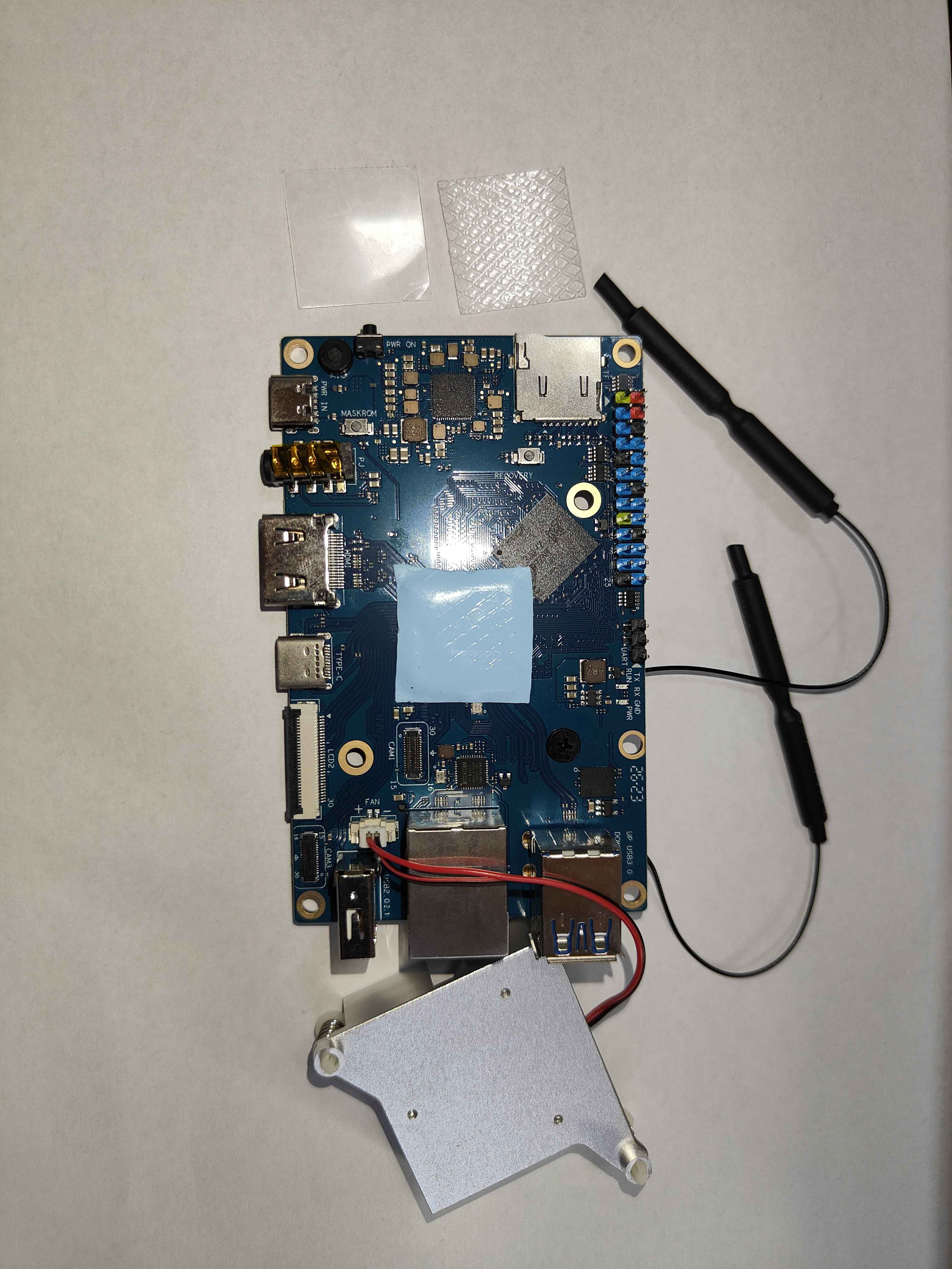 An Orange Pi 5 seen from above with a square blue thermal pad laid on the SoC in the middle of the board, the two peeled-off films above it, and the fan heatsink waiting upside down below the board with its lead already in the FAN socket