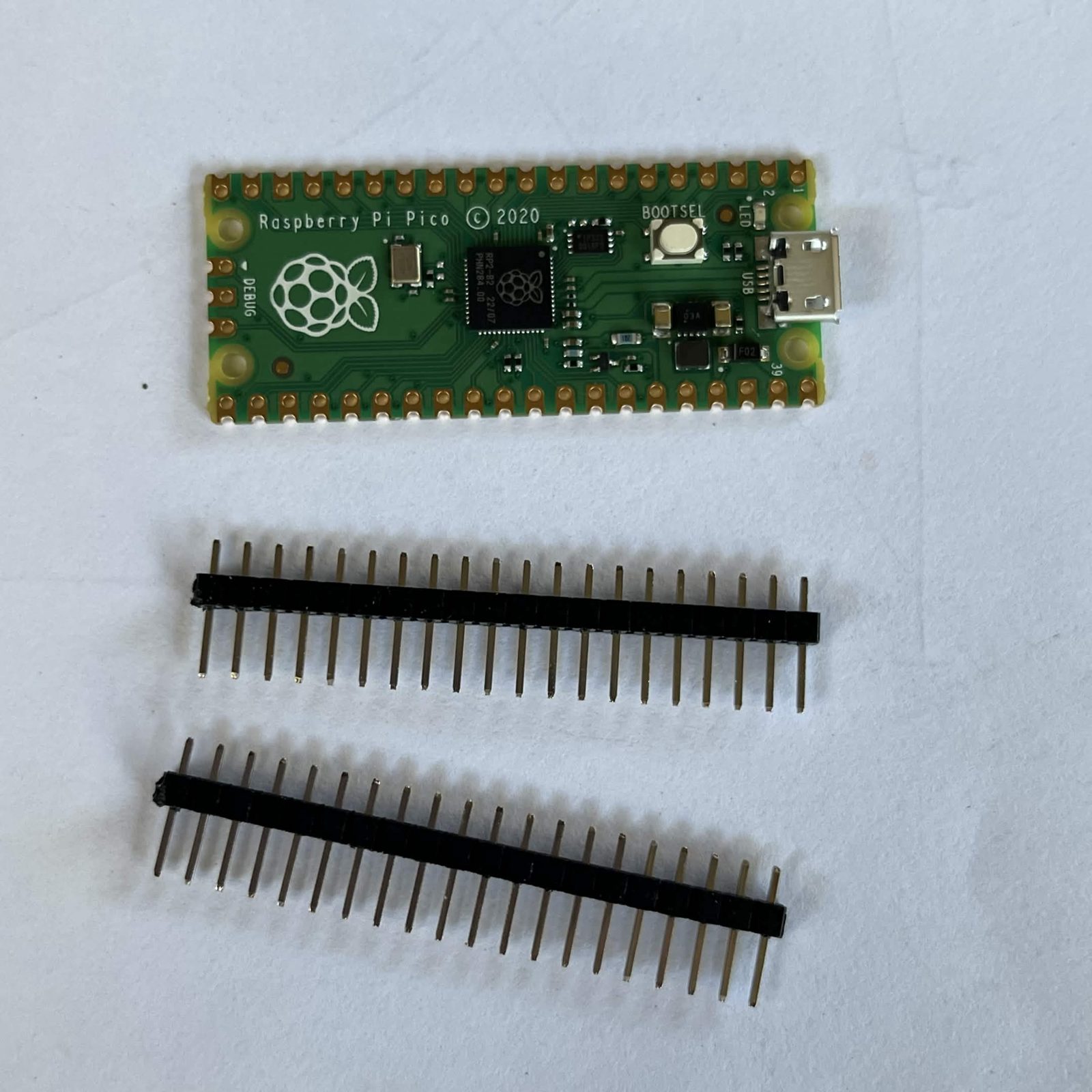 A bare Raspberry Pi Pico on a white surface with two broken-off rows of twenty black header pins lying below it