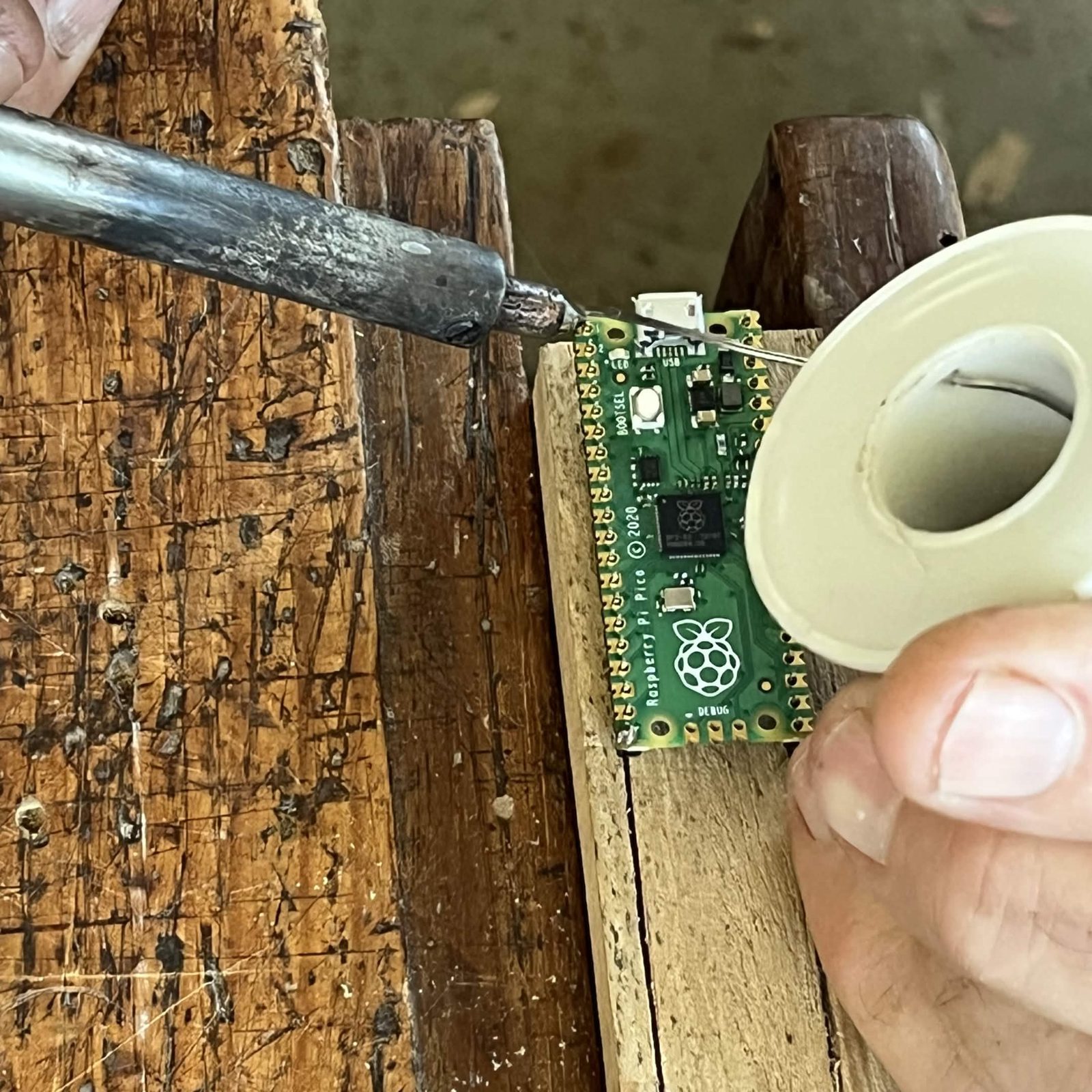 A soldering iron tip and a length of solder meeting a corner pin on the chip side of a Pico, with the pin at the far end of the same row already soldered, the board sitting on its wooden support in a vice