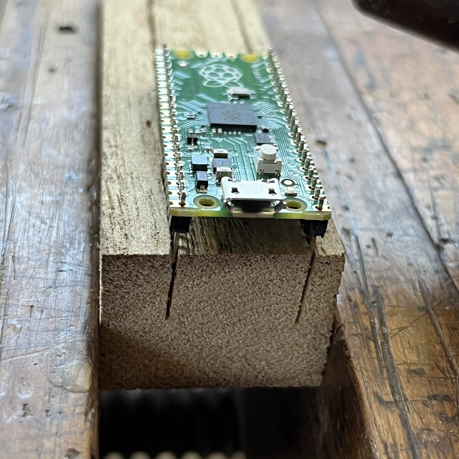 A Raspberry Pi Pico sitting flat on a block of soft wood, its two rows of pins dropped into two saw slots cut into the block