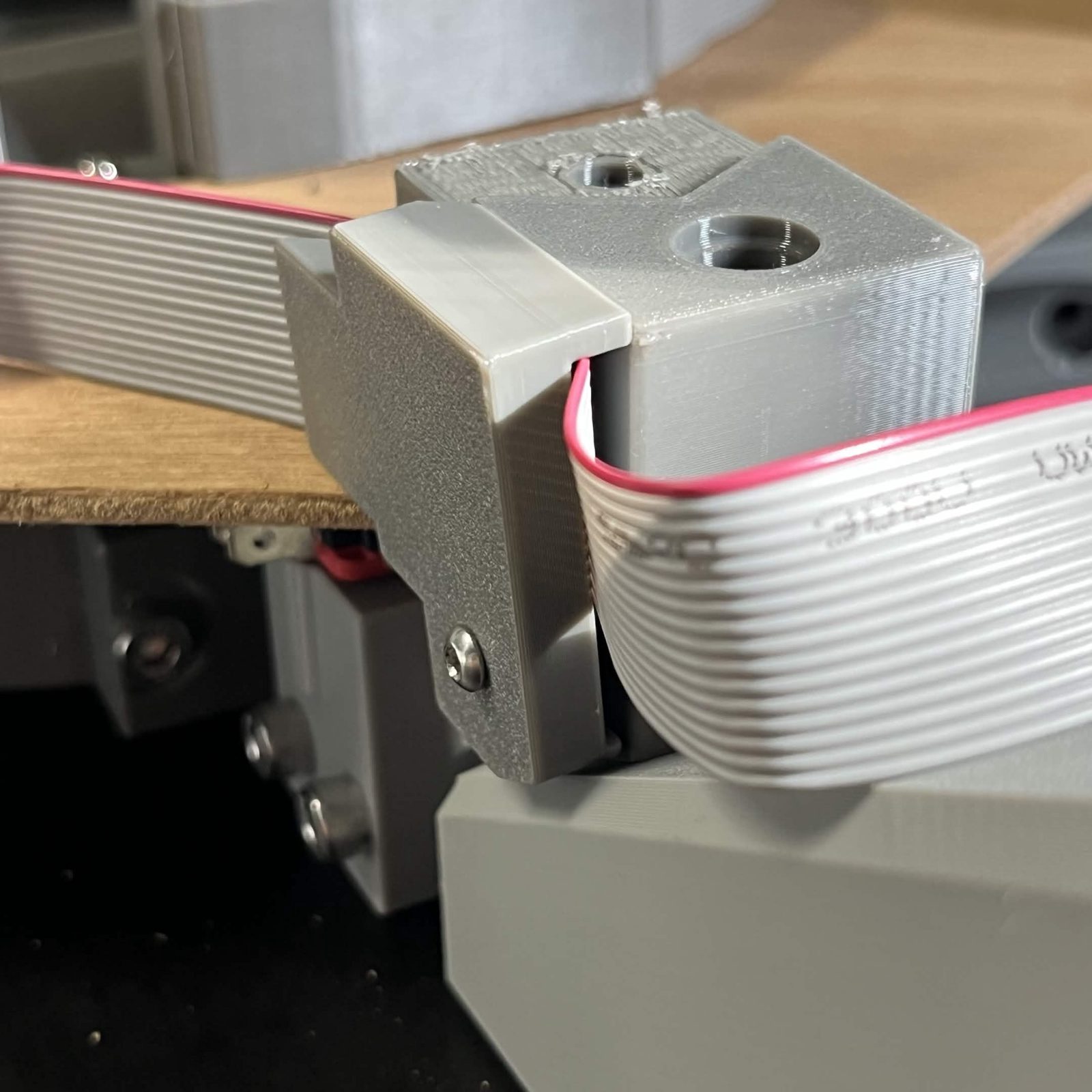 The ribbon cable running under the clamp on the bracket and folding back on itself, the red stripe along its edge, with one screw holding the clamp