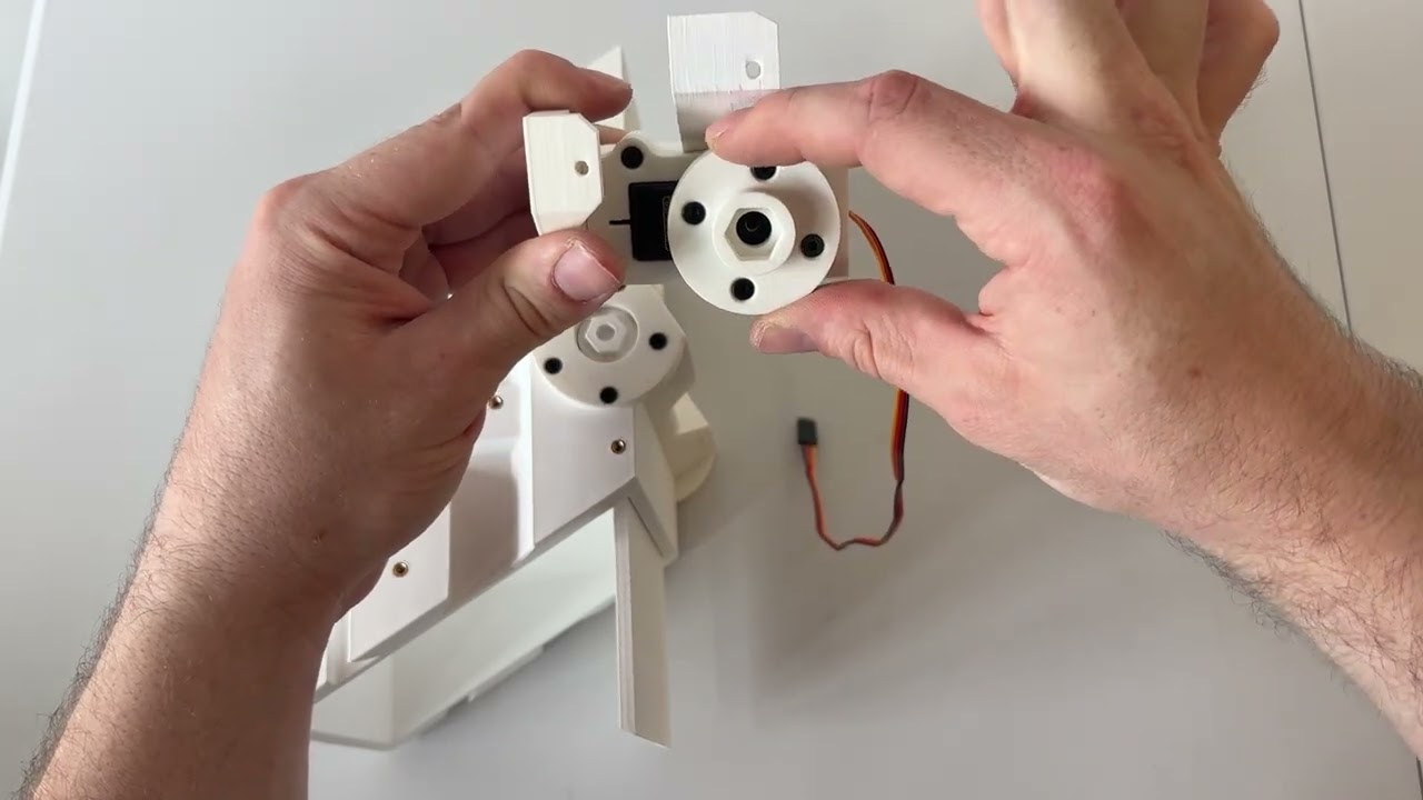 Photograph of a real door module part-way through assembly: a hand holds the assembled servo adapter, a white disc with a hexagonal boss in the middle and four countersunk screws around it, in front of the MG995 in its printed bracket