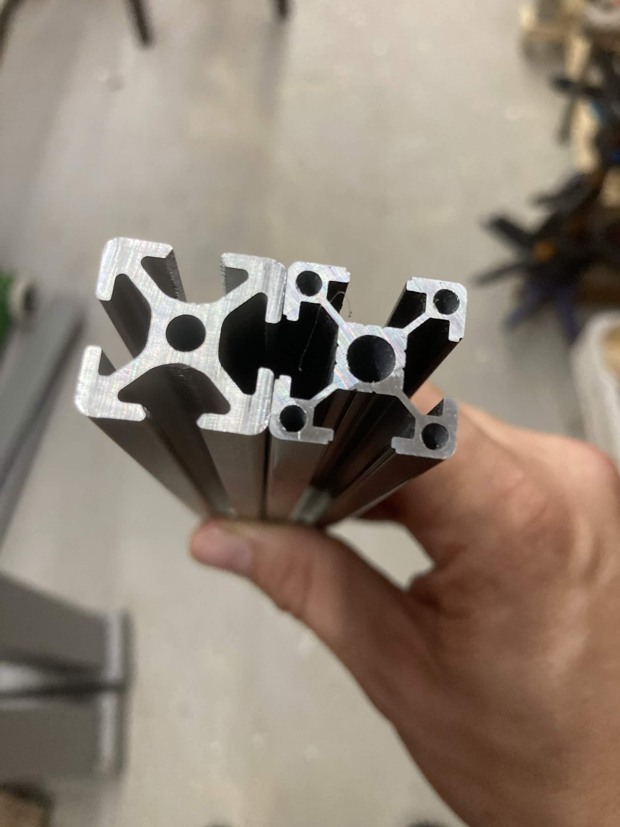 Two 2020 extrusion profiles held end-on side by side — the right one has angular T-slot corners, the left one has rounded corners